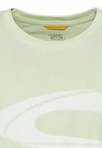 Lys grøn t-shirt med rund hals og subtil tekstur, der har et hvidt prikket spiral mønster over fronten. Mærkets label synlig indeni.