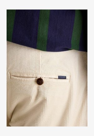 Cremfarbige Cordhosen mit strukturiertem Finish und einem braunen Knopfakzent, ausgestattet mit einem kleinen blauen "JOULES"-Label in der Nähe der Gesäßtasche.
