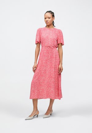 Rød midikjole med hvite polkadotter, korte puffermede ermer, innsvingt midje og flytende skjørt. Kombinert med lysegrå hælede sko.