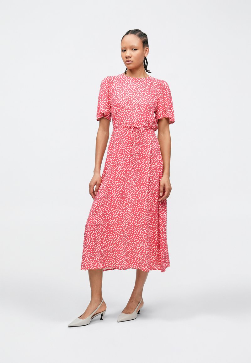 Κόκκινο midi φόρεμα με λευκές πουά βούλες, κοντά μανίκια με φουσκάλες, σφιχτή μέση και ρέον μανίκι. Συνδυασμένο με ανοιχτό γκρι παπούτσια με τακούνι.