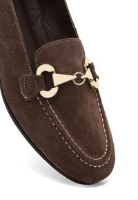 Loafer en suède marron avec un bout pointu, un accent en métal doré, et des détails de couture beige le long des bords.