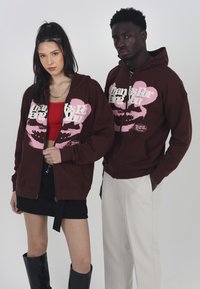 Braune Zip-Hoodies mit pinkfarbigem Grafiketext und Lippenabdruck. Kombiniert mit schwarzem Rock und Stiefeln; Modell links, Hose rechts.