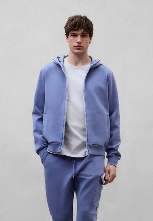 Joven con sudadera con capucha azul claro con cremallera, camiseta blanca y pantalones deportivos azul claro a juego, sobre un fondo liso.