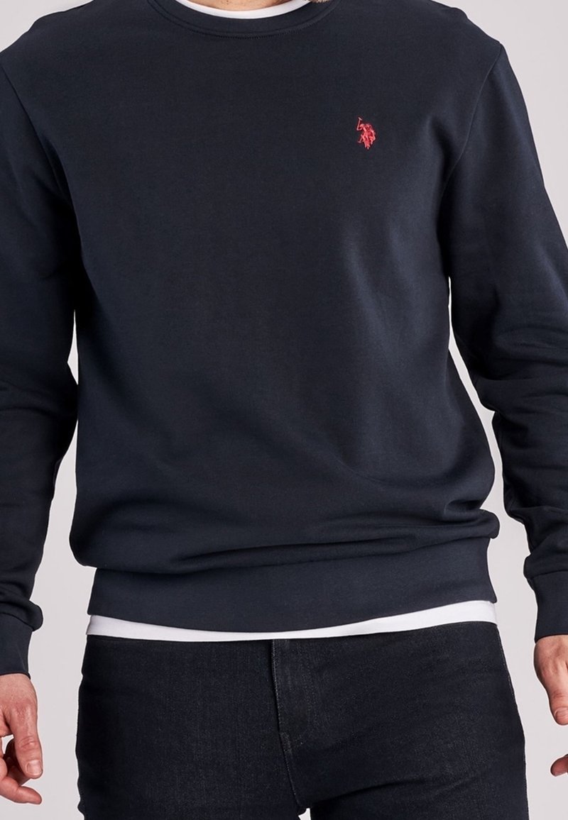 Mörkblå crewneck sweatshirt i mjukt material, med en liten röd logotyp på bröstet. Klippt med ribbade muddar och nederkant, i kombination med mörka jeans.
