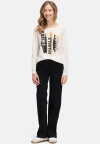 Kremowa koszula z długim rękawem z czarnym i pomarańczowym napisem „SMILE” i „SPARKLE”, w połączeniu z czarnymi spodniami i białymi trampkami. Gładka tkanina, luźny krój.