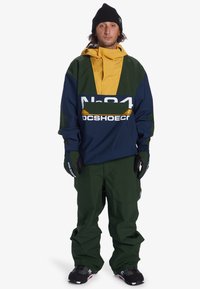 Color-blocked snowboarding outfit with navy blue, green, and yellow hues. Features a large logo, a hood, and baggy green pants.