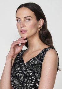 Haut sans manches noir à motifs floraux avec un décolleté en V ; présente un design complexe de motifs floraux blancs et verts. Boucles d'oreilles en créoles dorées.