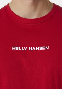 Červené bavlněné tričko s kulatým výstřihem, na němž je bílý vyšitý nápis "HELLY HANSEN" napříč hrudníkem. Hladký povrch.