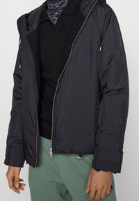 Sort polstret jakke med lynlås foran, hætte og elastiske manchetter. Fremstillet af glat, vandtæt materiale. Båret over en sort skjorte og grønne bukser.