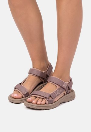 Füße in mauvefarbenen Sandalen mit offenem Zehenbereich und verstellbaren Klettverschlüssen auf beigen, strukturierten Sohlen vor weißem Hintergrund.