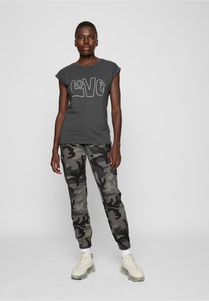 Egy nő áll, sötétszürke "LOVE" feliratú pólóban, szürke és fekete terepmintás nadrágban, fehér zokniban és bézs sportcipőben egy egyszínű háttér előtt.