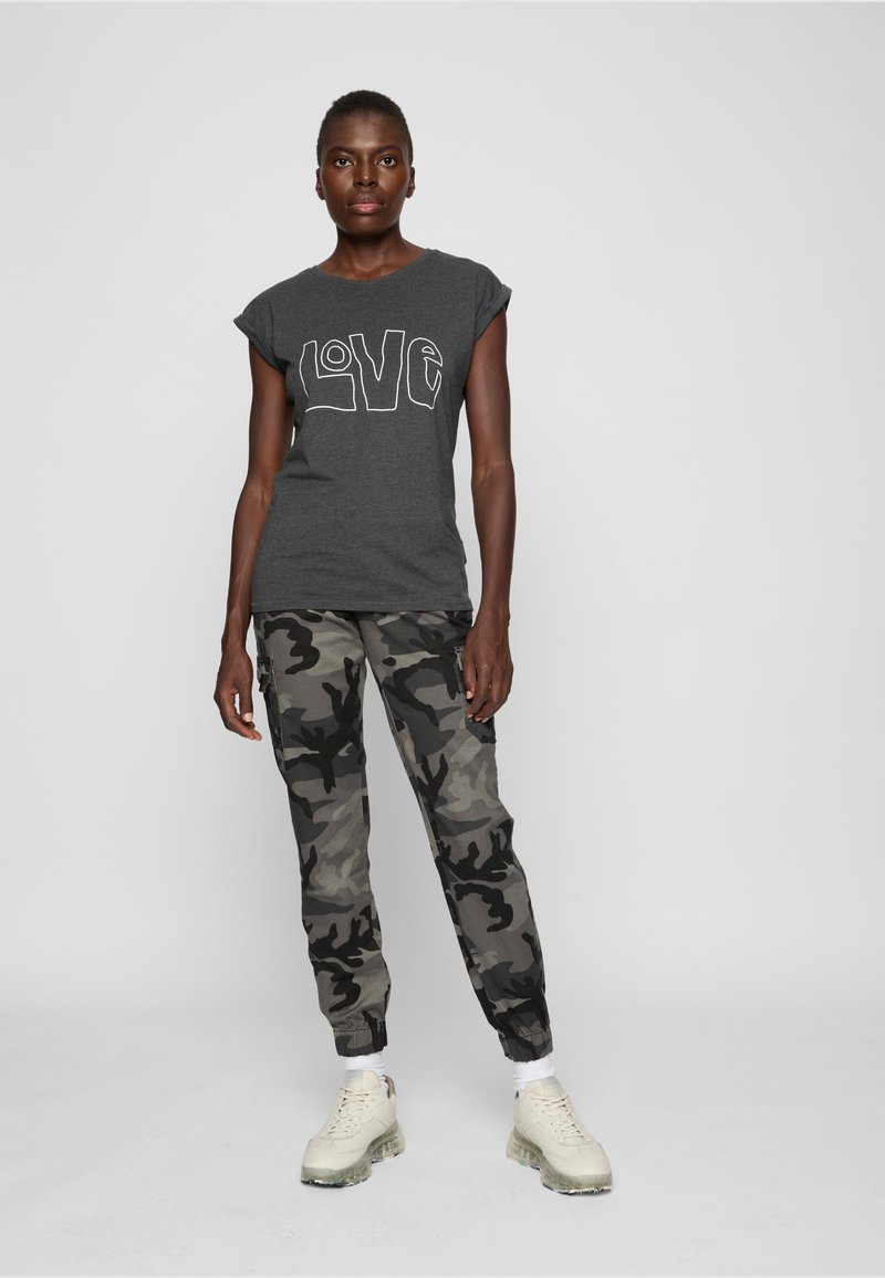 O femeie stă în picioare purtând un tricou gri închis cu inscripția „LOVE”, pantaloni de camuflaj gri și negri, șosete albe și adidași bej, pe un fundal simplu.