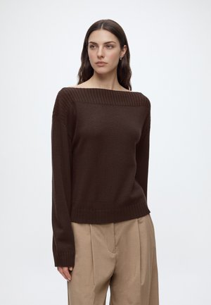 Femme aux longs cheveux foncés portant un pull marron foncé épaules dénudées et un pantalon plissé beige taille haute sur un fond uni.