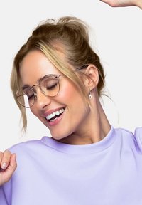 Runda, guldmetalliska glasögon; ljuslila topp med mjuk textur. Personen har bakåtrettet hår och bär en hängande örhänge med en klar accent.