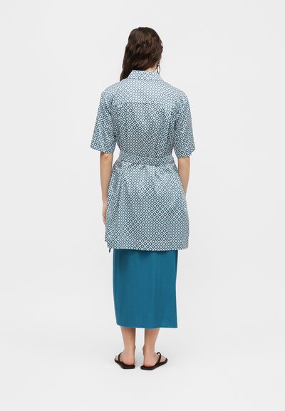 Γυναίκα που στέκεται με την πλάτη, φοράει μπλε τούνικα με γεωμετρικό σχέδιο και κοντά μανίκια πάνω από μια μονόχρωμη μπλε midi φούστα και μαύρα σανδάλια.