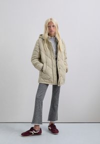 Jonge vrouw staat met haar handen in haar zakken, gekleed in een beige, gewatteerde jas met capuchon, een grijze trui, grijze broek met luipaardprint en bordeauxrode sneakers.