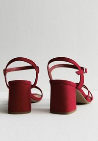 Sandali con tacco in suede rosso, con tacco a blocco, design aperto, cinturino alla caviglia e fasce minimaliste sulla parte superiore del piede. Superficie texturizzata e finitura liscia.