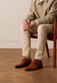 Braune Lederschuhe mit einem schlanken Design und einem Schnallenakzent. Glatte Textur und spitz zulaufende Form. Getragen mit hellen Hosen.