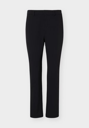 Persona con un polo beige de manga corta, pantalones negros con cinturón marrón y zapatillas blancas, de pie frente a un fondo claro y liso.