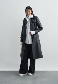 Long manteau en cuir noir avec des boutons, poches latérales et un design ajusté, porté sur une chemise blanche et associé à un pantalon noir large.