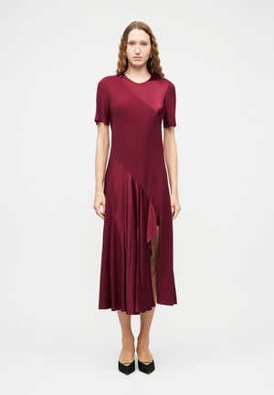 Mujer con cabello ondulado vistiendo un vestido midi asimétrico burdeos con mangas cortas y zapatos de tacón puntiagudos negros, posando frente a un fondo liso.