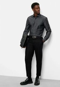 Donkergrijs overhemd met knopen, zwarte broek en zwarte schoenen. Een zwarte tas dragend; gladde stof met een getailleerd ontwerp. Casual zakelijke uitstraling.