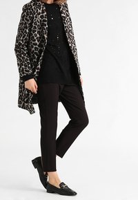 Persona vestida con pantalones negros, suéter negro perforado, abrigo de estampado de leopardo y mocasines negros con hebilla dorada, de pie contra un fondo blanco.