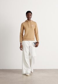Felpa con cappuccio beige con cordini e logo, abbinata a pantaloni larghi bianchi e sneaker metalliche, mentre tiene una pochette argentata.