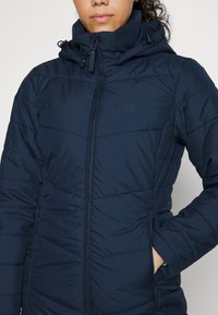 Tmavě modrá péřová bunda s vysokým límcem, předním zipem a vodorovným stehováním. Má dvě boční kapsy a nastavitelnou kapuci.