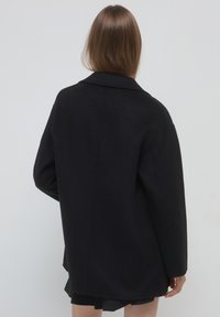 Cappotto di lana nera con vestibilità rilassata, colletto a punta e maniche lunghe. Presenta una texture liscia e una silhouette elegante.