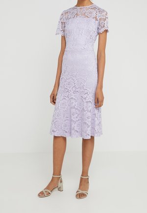 Kvinde iført en knælængde, kortærmet lavendel blonderkjole med blomsterprint og beige blokhælssandaler med åbne tæer.