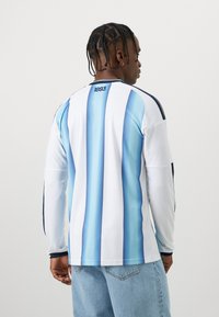 Camisola de desporto de mangas longas em branco com riscas verticais azuis. Apresenta um decote redondo e detalhes em azul-marinho nos ombros. As costas exibem "1893."