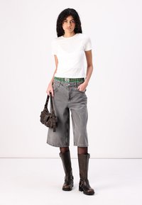 T-shirt bianco aderente, pantaloni corti in denim grigi a gamba larga con cintura a quadri verdi, stivali marroni al ginocchio e una piccola borsa marrone.