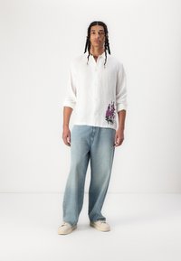 White linen long-sleeve shirt with floral embroidery in purple, paired with relaxed-fit light blue jeans and beige sneakers.