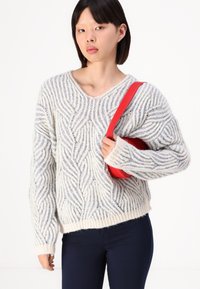 Crème en blauwe gebreide trui met een diepe V-hals en geribbelde manchetten. Model houdt een rode tas vast, draagt marineblauwe broek.