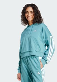 Teal cropped huppari, jossa on etutasku, nyörillä varustettu huppu ja vaaleanpunainen Adidas-logo. Hihoissa on valkoiset raidat. Pehmeä kangas.