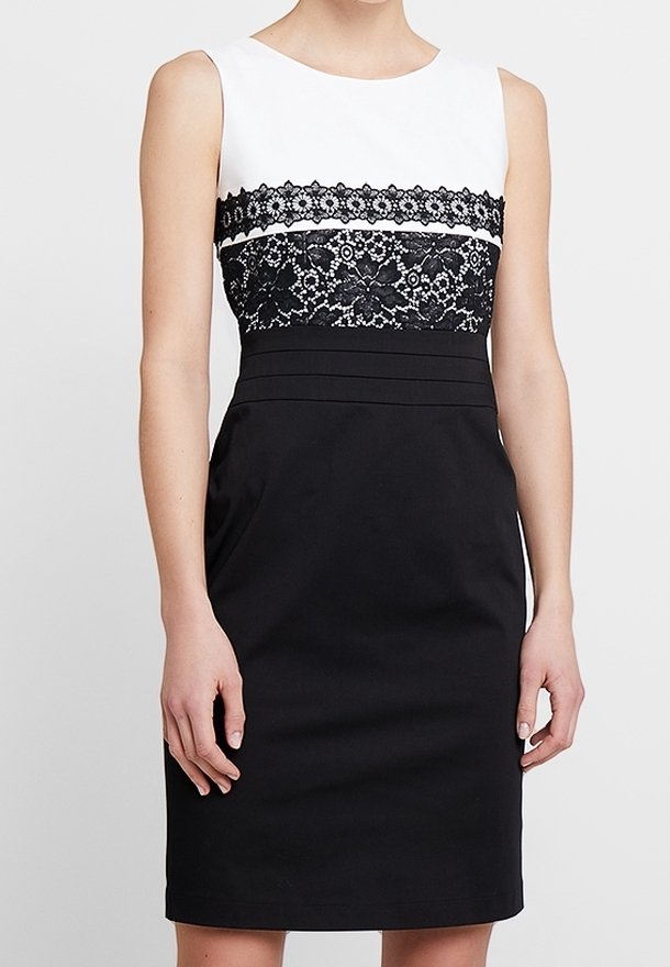 Robe noire et blanche sans manches avec un détail en dentelle florale sur la poitrine et une taille cintrée, portée par une personne debout.