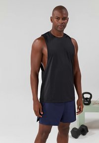 T-shirt de sport sans manches noir en tissu lisse, assorti à des shorts bleu marine. Design épuré, coupe ample, idéal pour les entraînements. Équipement de fitness en arrière-plan.