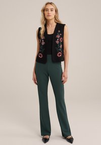 Juodas pagalvėlės formos liemenė su išsiuvinėtais gėlės raštais, dėvima ant juodo marškinėlio ir derinama su žaliais siaurais džinsais bei juodais aštriakampiais bateliais.