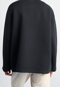 Svart sweatshirt med en lös passform, rund halsringning och långa ärmar. Slät textur med minimal logga på baksidan. Matchad med ljusa byxor.