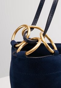 Sac à main en daim bleu marine avec des sangles en cuir noir passant à travers de grandes anneaux métalliques dorés sur le bord supérieur.