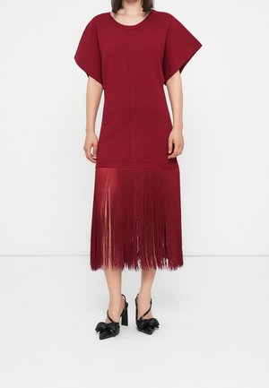 Femme portant une robe bordeaux longueur genoux avec des manches courtes larges et des franges en bas, associée à des chaussures à talons noires.