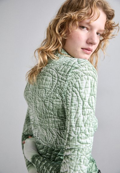 Quilted jacket in light green fabric, featuring a textured floral pattern. The design has a fitted silhouette and high collar.