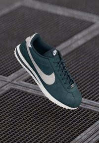 Zelene sportske cipele od zamšene kože s sivim akcentima, koje imaju bijeli Swoosh logo, teksturiranu podlogu i dizajn s vezanjem.