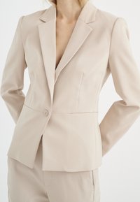 Bežový přizpůsobený blejzr s jedním knoflíkem, s klopami a strukturovaným střihem. Hladká textura s jemnými švy.