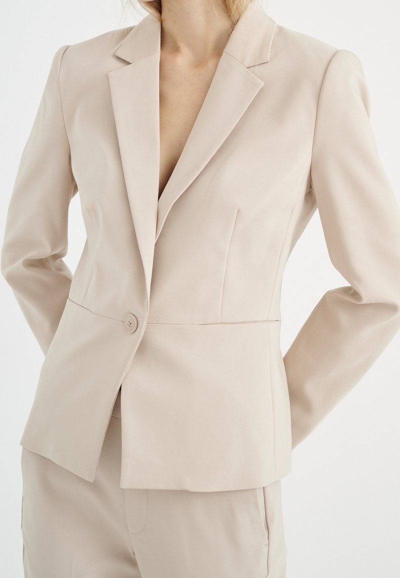 Bežový přizpůsobený blejzr s jedním knoflíkem, s klopami a strukturovaným střihem. Hladká textura s jemnými švy.