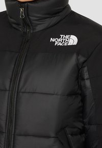 Chaqueta de plumas negra con un alto cuello, que presenta un cierre de cremallera plateado y un logo bordado en el pecho. Textura suave con diseño acolchado.