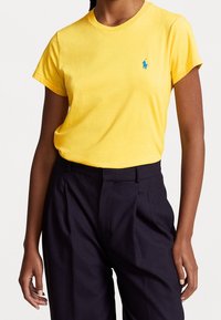 Žena v jasně žlutém krátkém tričku zasunutém do vysokých černých kalhot se sklady stojí na bílém pozadí.