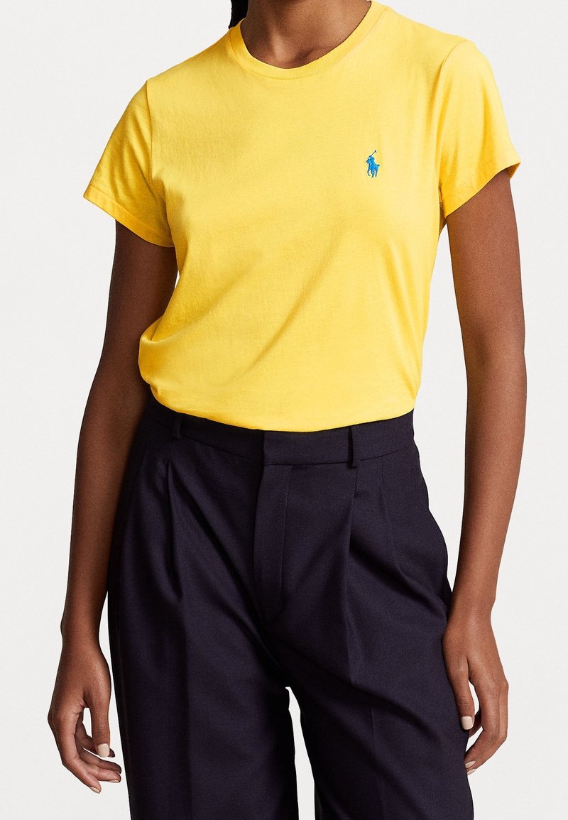 Žena v jasně žlutém krátkém tričku zasunutém do vysokých černých kalhot se sklady stojí na bílém pozadí.