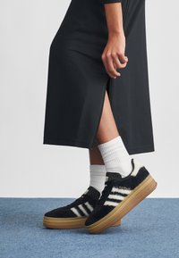 Sapatilhas Adidas Gazelle pretas felpudas com três riscas brancas, sola de borracha castanha e parte superior texturada. Usadas com meias brancas até ao tornozelo sobre um tapete azul.